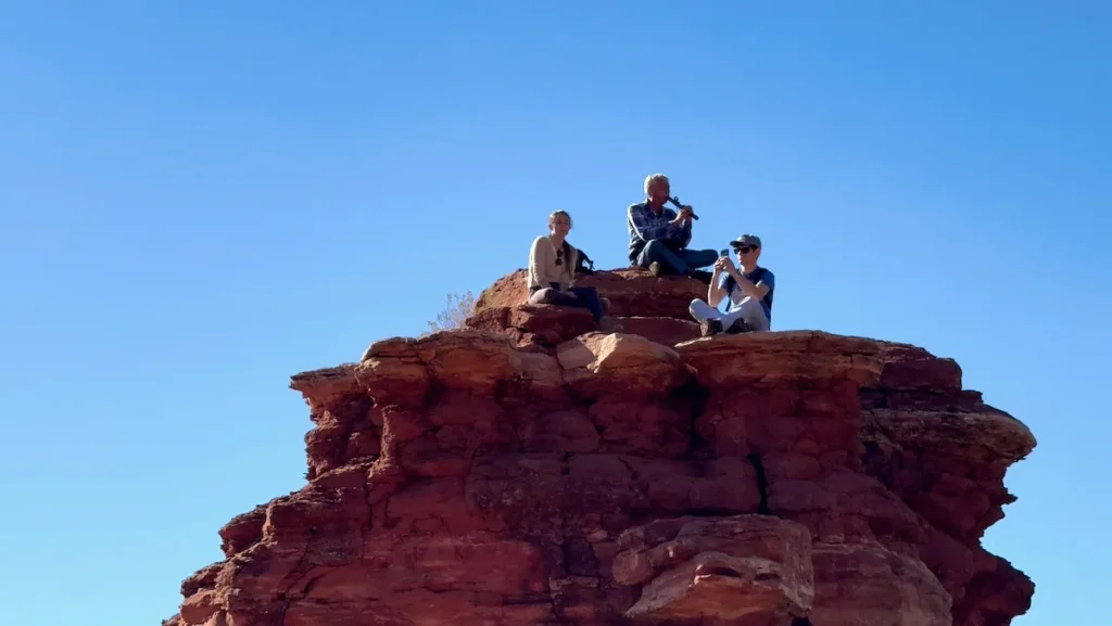 A flute player sitting on a red rock ledge in Boynton Canyon, with others listening nearby.
