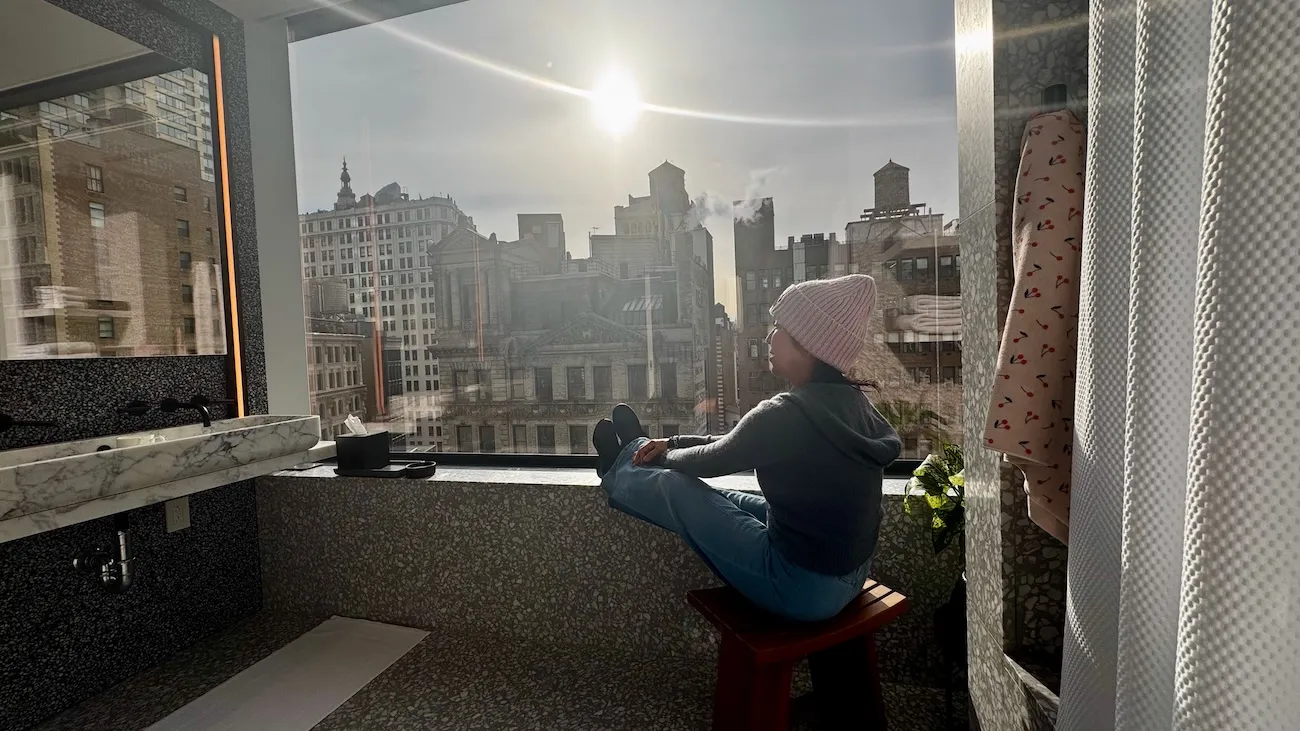 Woman sitting by a bathroom hotel window seat overlooking New York City buildings in the morning light at The Ritz-Carlton NoMad